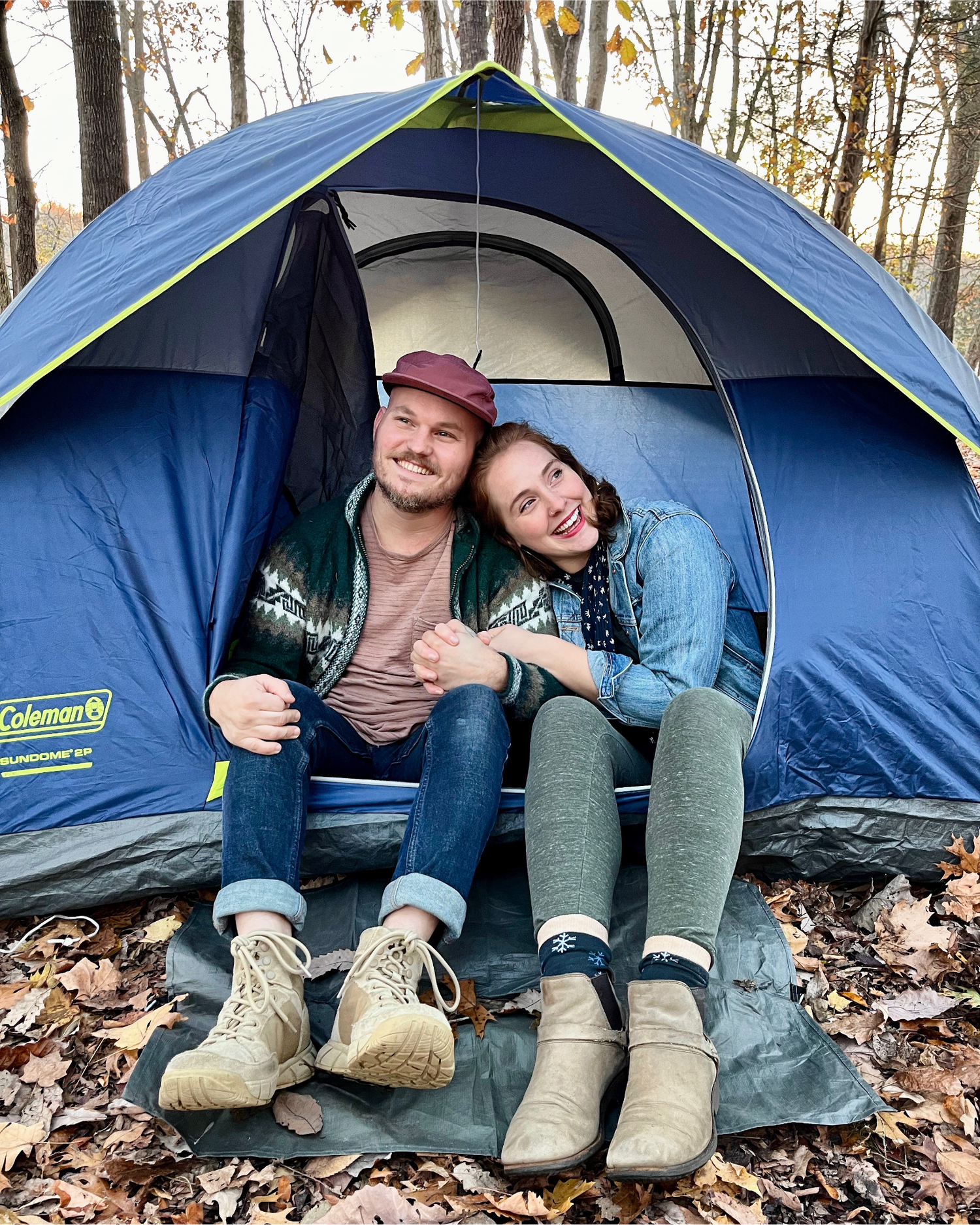 Two people sitting inside a blue Coleman tent in a forest setting.
